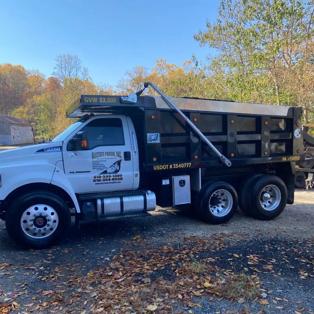 White and black Allstate Paving of PA dump truck with company lettering on the door, parked on gravel among autumn trees.
