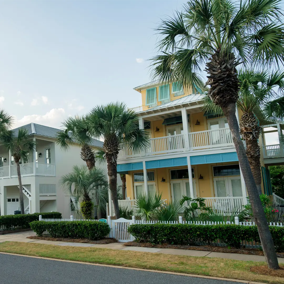 Smooth asphalt residential street lined with palm trees and multi-story coastal homes with porches and a picket fence.